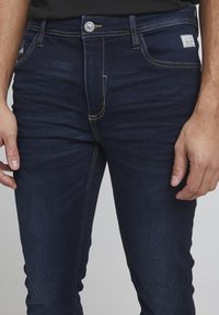 Jeans de mezclilla azul oscuro con costuras amarillas, que cuentan con una cremallera y un cierre de botón, bolsillos laterales y una pequeña etiqueta de la marca en la cinturilla.
