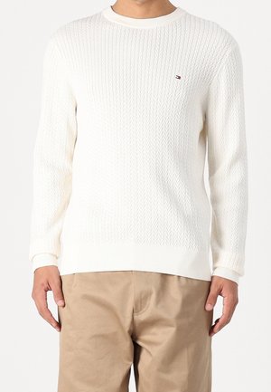 Homme portant un pull blanc texturé à col rond et un pantalon beige, debout avec les mains détendues le long du corps, fond neutre.