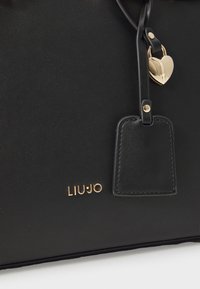Zwarte leren handtas met een gouden logo, hartvormige hardware en een rechthoekig label met stikdetails. Gladde textuur en een minimalistisch ontwerp.