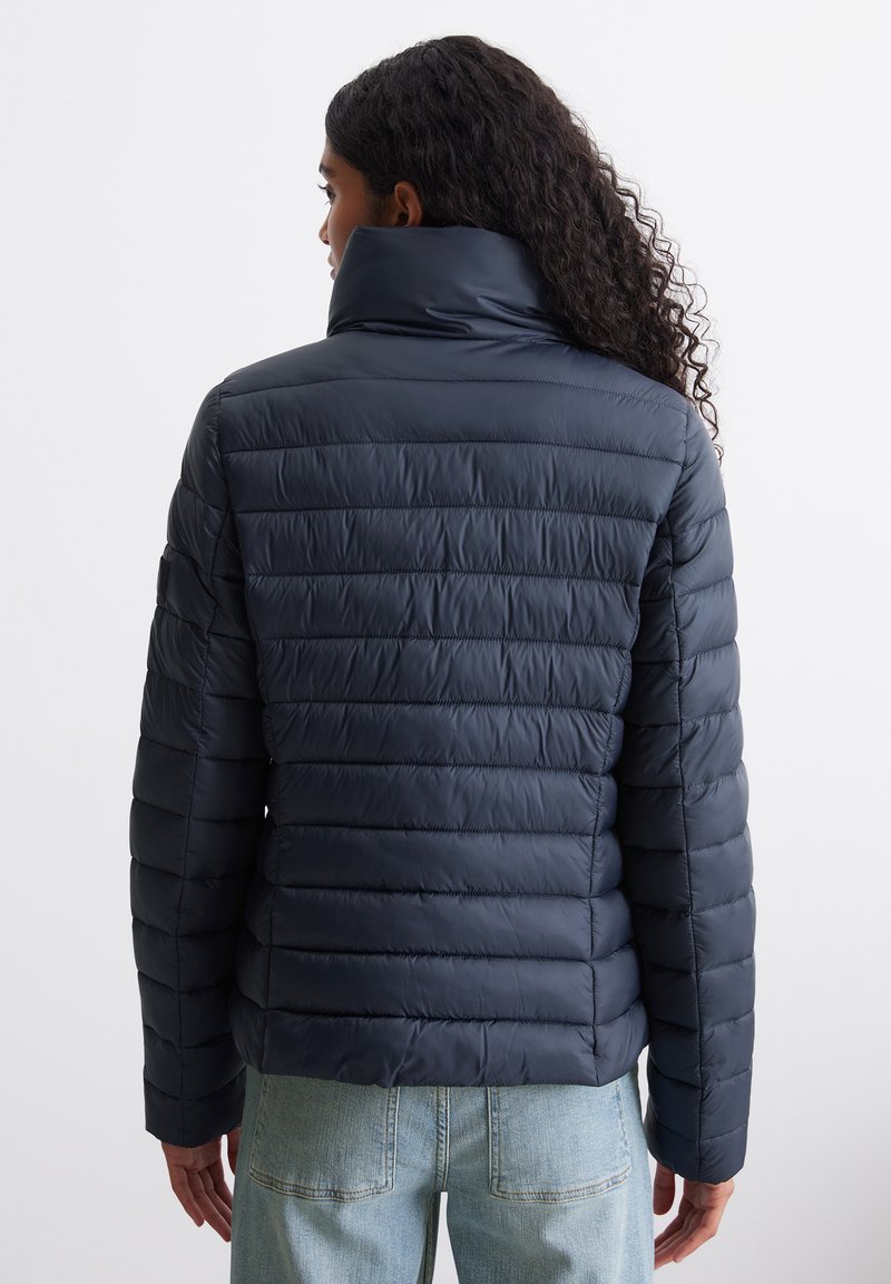 Marineblaue Steppjacke mit hohem Kragen, horizontalem Steppmuster, glatter Textur und langen Ärmeln, getragen über hellblauen Jeans.