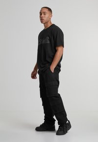 Tricou negru supradimensionat cu text gri, asortat cu pantaloni cargo negri prevăzuți cu mai multe buzunare și teniși negri înalți pe un fundal deschis.