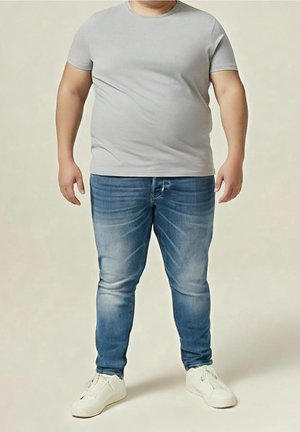 Homme portant un t-shirt gris clair, un jean bleu et des baskets blanches, debout devant un fond neutre clair.