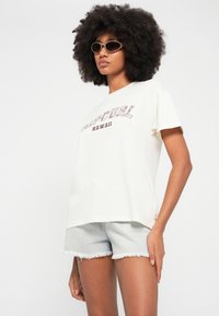 Roomkleurig T-shirt met korte mouwen en "Hawaii"-print, gedragen met lichte denim shorts. Model draagt ook ovale zonnebril en heeft textuurrijk krullend haar.