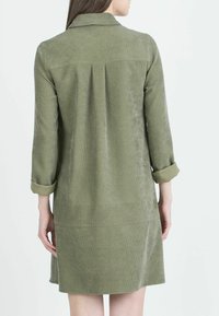Abito in corda verde oliva con colletto, maniche lunghe e polsini arrotolati. Presenta un dettaglio a pieghe sul retro e una silhouette dritta.