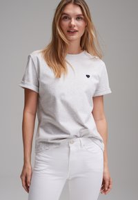 Graues Baumwoll-T-Shirt mit kurzen Ärmeln, rundem Halsausschnitt und einem kleinen schwarzen Herz, das auf der linken Brust gestickt ist. Kombiniert mit weißen Jeans.