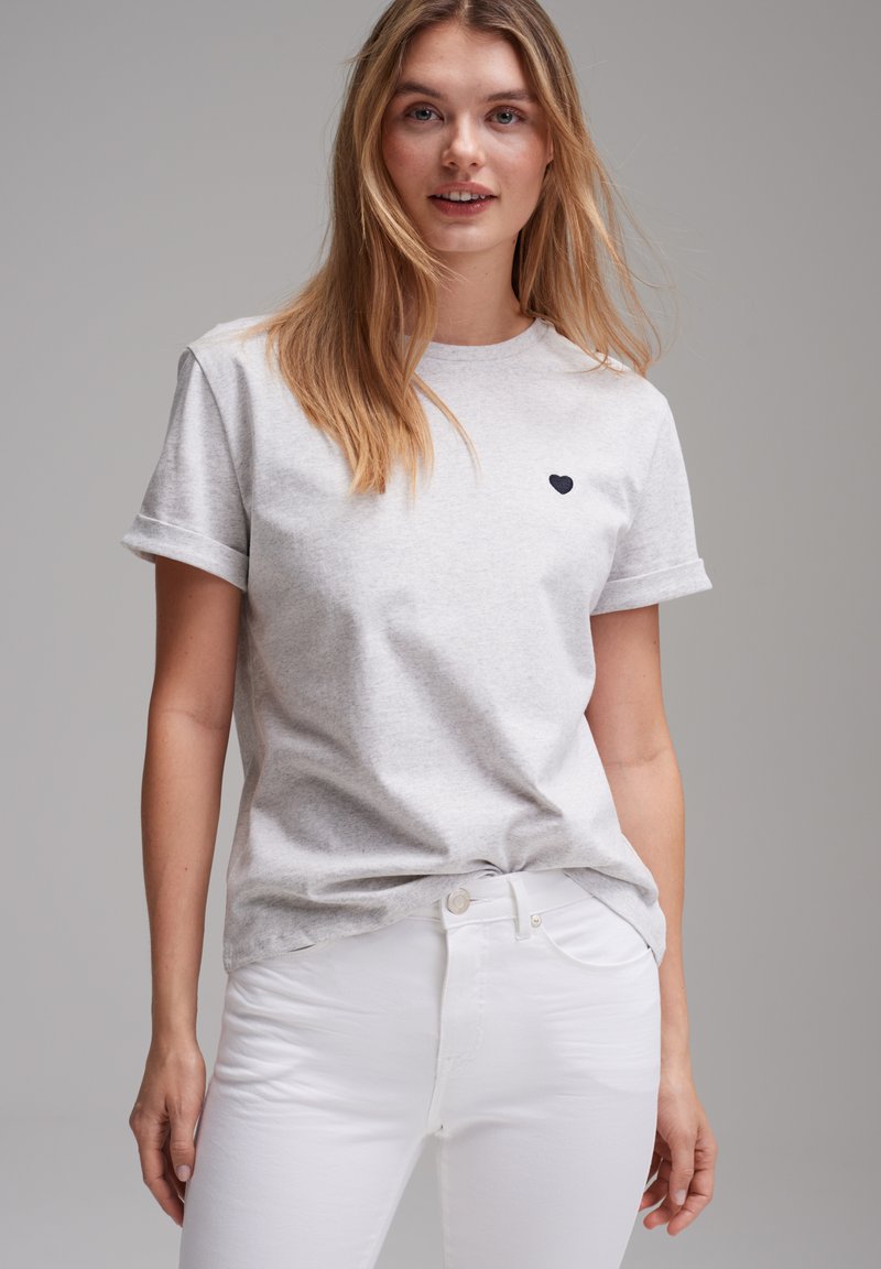Graues Baumwoll-T-Shirt mit kurzen Ärmeln, rundem Halsausschnitt und einem kleinen schwarzen Herz, das auf der linken Brust gestickt ist. Kombiniert mit weißen Jeans.