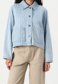 Ljusblå denimjacka med vertikala vita ränder, knappstängning och två framfickor. Har en klassisk krage och avslappnad passform.