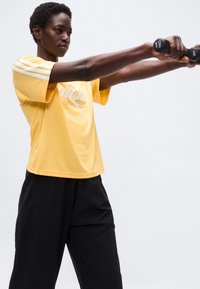 Άτομο με κίτρινη μπλούζα Adidas και μαύρο παντελόνι κρατά δύο αλτήρες 1 κιλού ευθεία μπροστά, ασκούμενο μπροστά σε απλό φόντο.