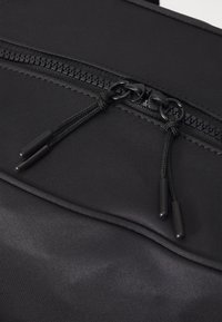 Black fabric backpack with smooth texture, featuring a centered zipper and two black cord pulls with rubber tips for easy handling.