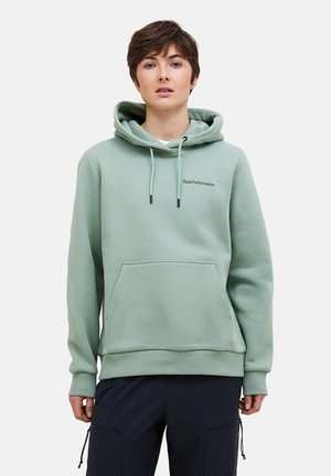 Personne portant un sweat à capuche PeakPerformance vert clair avec une poche devant et un pantalon foncé, debout devant un fond uni.