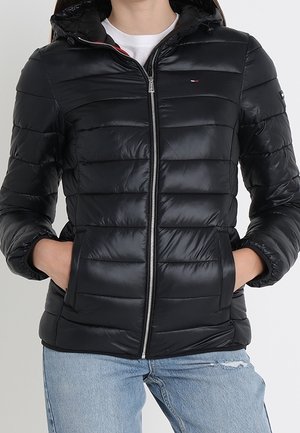 Person iført en sort quiltet dunjakke med frontlynlås og hænder i lommerne, kombineret med lyseblå revnede jeans.