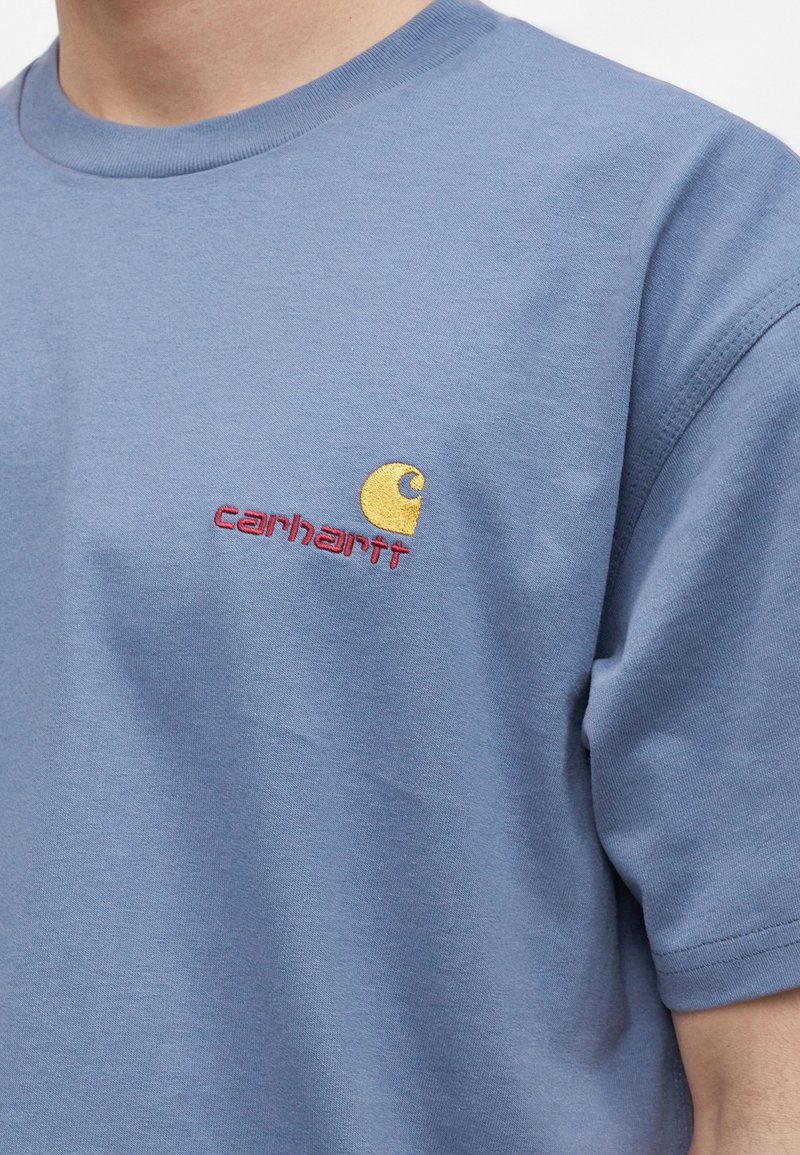 Blue short-sleeve shirt with yellow and red Carhartt logo embroidered on the left chest, worn by a person.