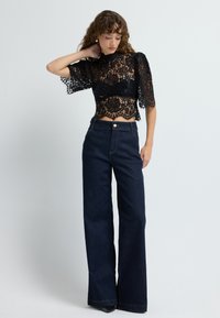 Top in pizzo nero con maniche a sbuffo corte e orlo scallop abbinato a jeans a vita alta blu scuro a gamba larga con chiusura anteriore a bottone.