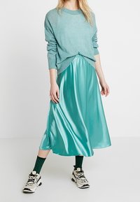 Světle modrý svetr přes lesklou tyrkysovou saténovou midi sukni. Outfit doplňují zelené ponožky s motivem a bílé tenisky.