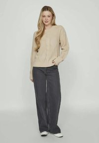 Μπεζ πουλόβερ σε κοτλέ ύφανση με V-λαιμό και ριχτές μανσέτες, συνδυασμένο με φαρδιά σκούρα γκρι τζιν και λευκά αθλητικά παπούτσια. Μαλακή υφή, χαλαρή εφαρμογή.