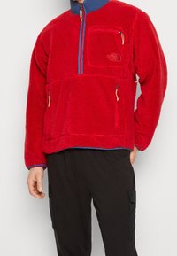 Sudadera de polar roja con cremallera de un cuarto, cuello azul, con un bolsillo frontal y cremalleras laterales. Textura suave con un tacto cálido.