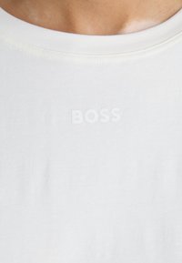 T-shirt en coton blanc avec un col rond. Présente un logo "BOSS" en relief de la même couleur blanche. Texture douce et design minimaliste.