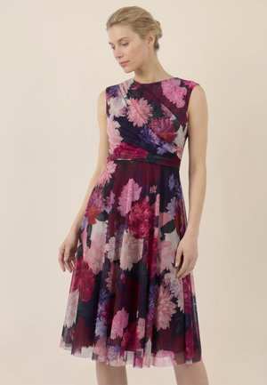 Frau in ärmellosem, knielangem Kleid mit lila, rosa und rotem Blumenmuster, stehend vor einem schlichten beigen Hintergrund.