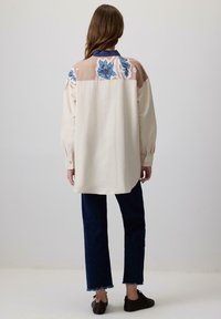 Lichtbeige oversized shirt met blauwe bloembroderie op een tan schouderstuk, gecombineerd met donkerdenim broek en bruine schoenen. Lange mouwen, knopen bij de manchetten.