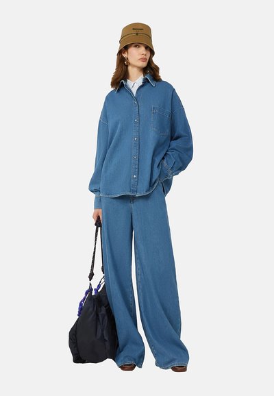 Donna che indossa una camicia di jeans blu larga e jeans a gamba larga, cappello bucket marrone, tiene una borsa nera con manici di corda, in piedi su sfondo bianco.