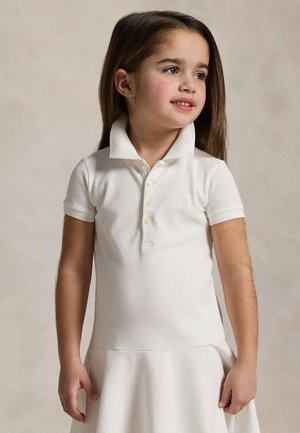 Jeune fille aux longs cheveux bruns portant une robe blanche à manches courtes avec col, regardant sur le côté devant un fond neutre uni.