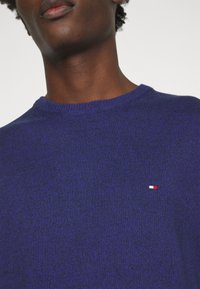 Gros plan sur une personne portant un pull en tricot bleu avec un petit logo rectangulaire rouge, blanc et bleu sur la poitrine.