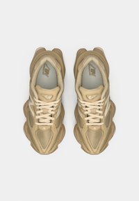 Chaussures de sport beiges avec des textures superposées, dotées d'une languette rembourrée, de lacets et d'un design de semelle incurvée. Logo New Balance de chaque côté.