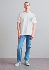 Camiseta blanca de algodón con un diseño abstracto azul en el lado izquierdo del pecho, combinada con jeans de denim azul claro y zapatillas grises.