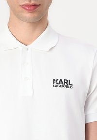 Valkoinen polo-paita, joka on valmistettu puuvillasta, ja siinä on klassinen kaulus sekä kolmesta nappia koostuva napitus. Rinnassa on musta "KARL LAGERFELD" -logo.