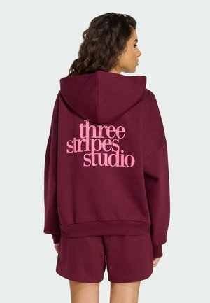 Felpa con cappuccio color marrone amaranto con una grafica rosa sul retro con la scritta "three stripes studio". Presenta un cappuccio con coulisse e polsini a costine.