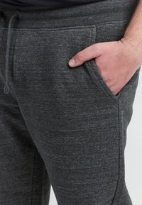 Szürke jogger nadrág texturált anyagból, oldalzsebekkel és húzózsinóros derékrésszel, közelről a zsebben lévő kézzel.