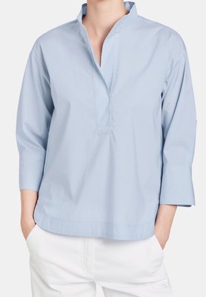 Chemise bleu clair à manches longues avec col montant et encolure en V ouverte, portée avec un pantalon blanc, mains dans les poches, visible des épaules jusqu’aux hanches.