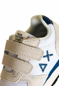 Sneaker bianco con accenti in suede beige, cinturini in velcro e simboli ricamati blu. Presenta un tessuto testurizzato e dettagli cuciti.