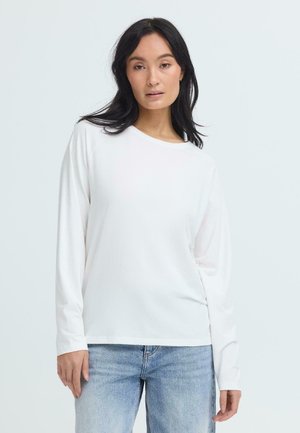 Vrouw met zwart haar, gekleed in een effen wit shirt met lange mouwen en lichtblauwe spijkerbroek, staande tegen een lichtgrijze achtergrond.