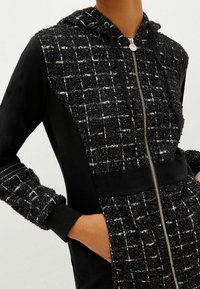 Chaqueta con capucha que presenta un tejido texturizado en negro y blanco, mangas y parte inferior en negro, con un cierre frontal y bolsillos laterales.