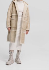 Beige faux shearling jakke med hætte, der har fire forlommer og kontrastfarvet kant. Sammen med en ribbet hvid kjole og taupe snørebåndsstøvler.
