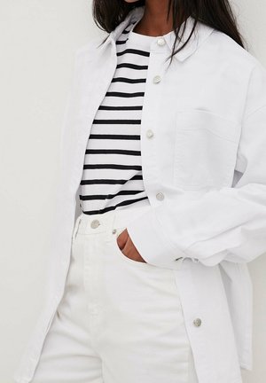 Une personne porte une chemise blanche boutonnée par-dessus un haut rayé noir et blanc, avec un pantalon blanc et une main dans la poche.