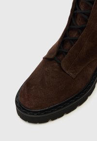 Gros plan sur une botte en daim marron foncé avec des lacets noirs et une semelle en caoutchouc noir épaisse sur fond blanc.