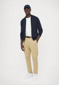 Námornícky modrý rebrovaný sveter cez biele tričko, khaki cargo nohavice a biele tenisky. Zodpovedajúca námornícka čiapka s logom. Prezentácia ležérneho outfitu.