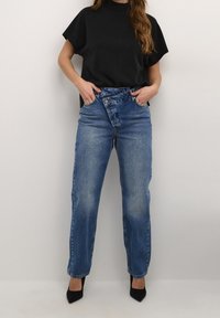 Kvinna som bär blå högmidjade jeans med asymmetrisk knappdetalj, svart kortärmad topp och svarta spetsiga klackar, med händerna i fickorna.