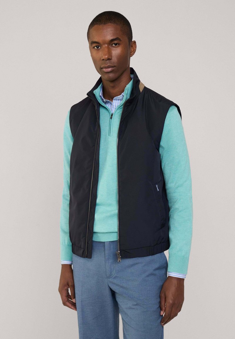 Granatowa kamizelka bez rękawów na jasnym swetrze w kolorze teal, z zamkiem z przodu i wysokim kołnierzem. Widoczne mankiety koszuli w niebiesko-białe paski.