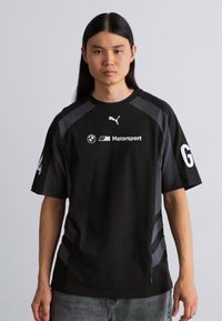 Sort kortærmet t-shirt med grå accenter, prydet med Puma- og BMW Motorsport-logoer. Inkluderer en buet kant og kontrasterende striber på skuldrene.