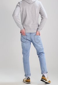 Felpa con cappuccio grigia, jeans azzurri chiari e sneakers marroni e nere. I jeans hanno una vestibilità rilassata e risvolti, con le tasche posteriori visibili.