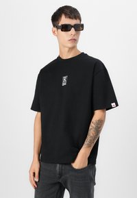 Black cotton t-shirt with short sleeves, featuring a small white logo on the chest. Model wears black square sunglasses.