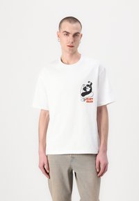 Camiseta blanca de algodón con un diseño gráfico negro en la zona izquierda del pecho que presenta el texto "HEART BASED" en naranja. Mangas cortas, corte relajado.