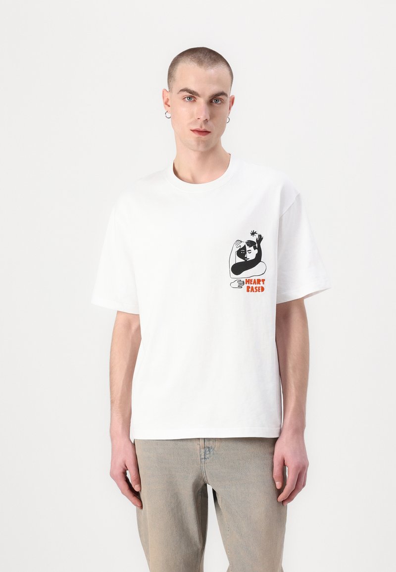 Camiseta blanca de algodón con un diseño gráfico negro en la zona izquierda del pecho que presenta el texto "HEART BASED" en naranja. Mangas cortas, corte relajado.