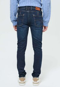 Jean bleu foncé à coupe slim, avec poches arrière et couture orange. Porté avec un pull en laine torsadée bleu clair et des baskets blanches.