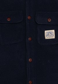 Navy fleece shirt with two chest pockets, brown snap buttons, and a woven label featuring a mountain graphic. Soft texture, casual design.