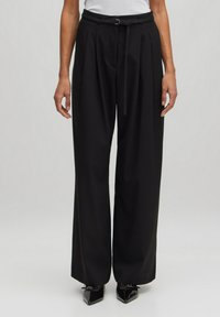 Schwarze weite Hosen mit hoher Taille, gefaltetem Vorderteil und passendem Gürtel. Der Stoff hat eine glatte Textur. Getragen mit spitzen schwarzen Schuhen.