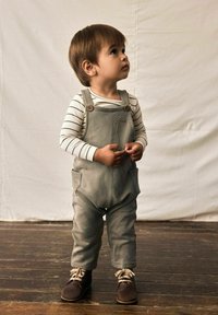 Overoles de algodón gris con dos bolsillos frontales, botones de madera, combinados con una camiseta de rayas blanca y gris de manga larga y zapatos marrones.